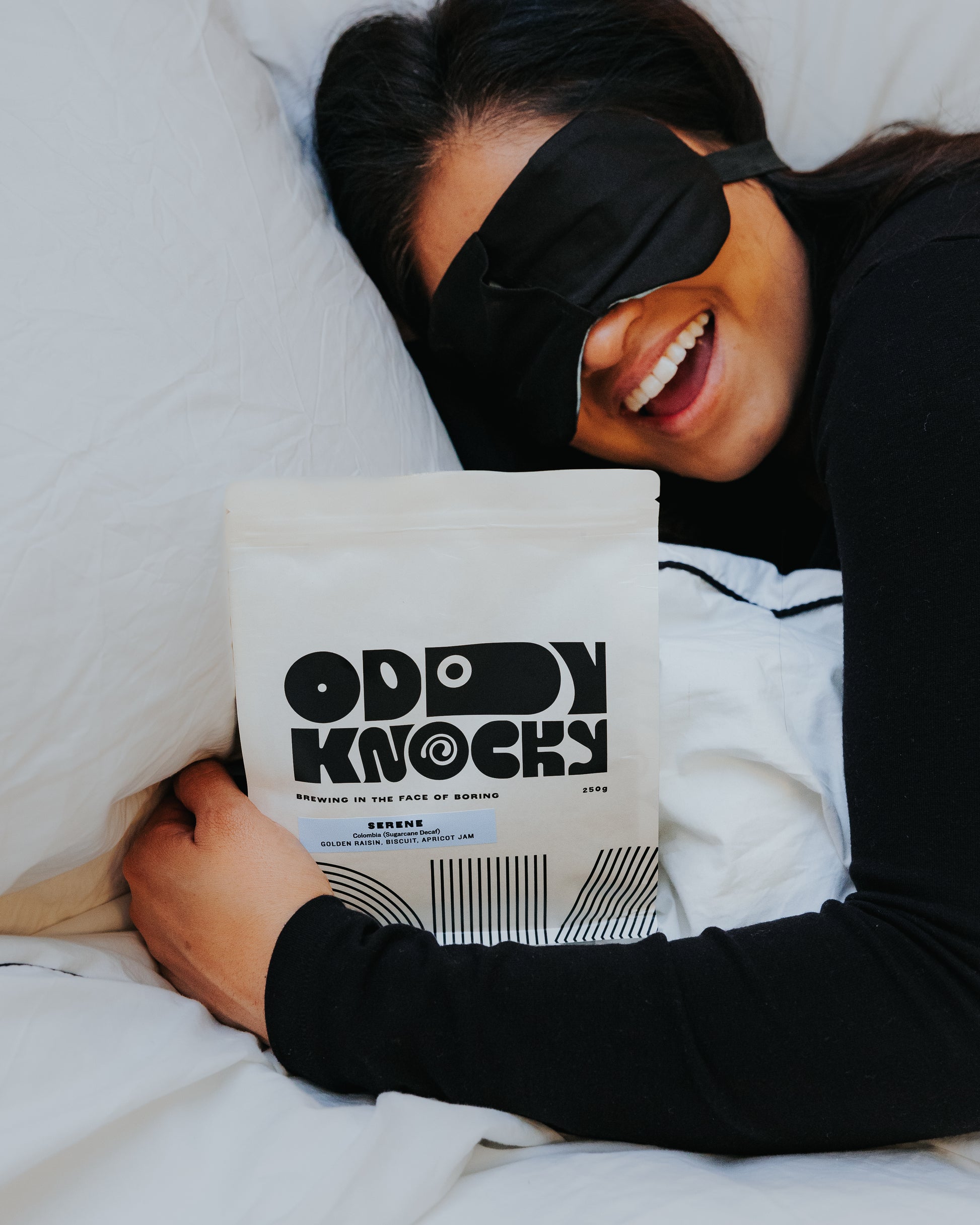 Person wearing a sleep mask, happily holding a bag of decaf coffee titled 'Serene (Decaf) – Colombia'.