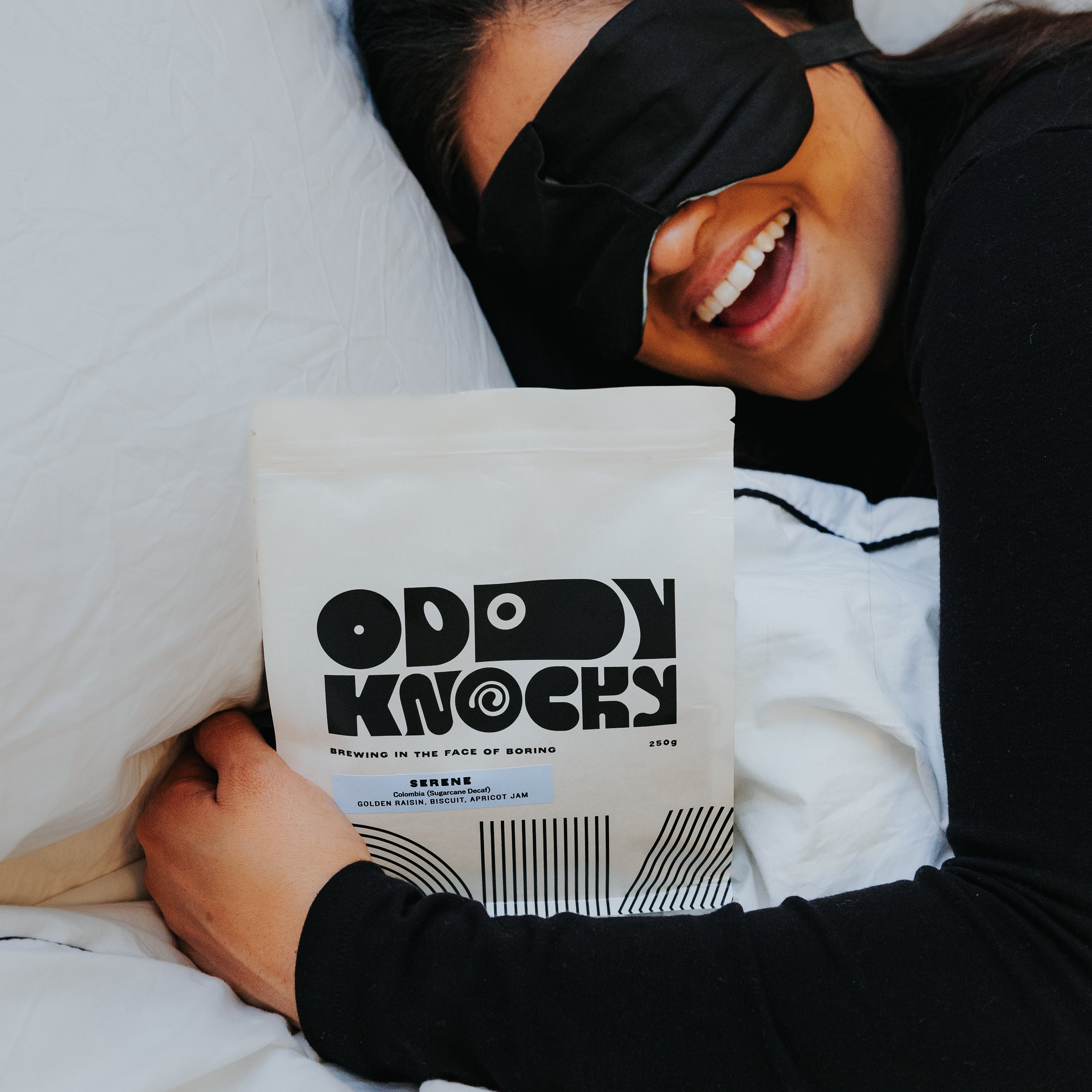 Person wearing a sleep mask, happily holding a bag of decaf coffee titled 'Serene (Decaf) – Colombia'.
