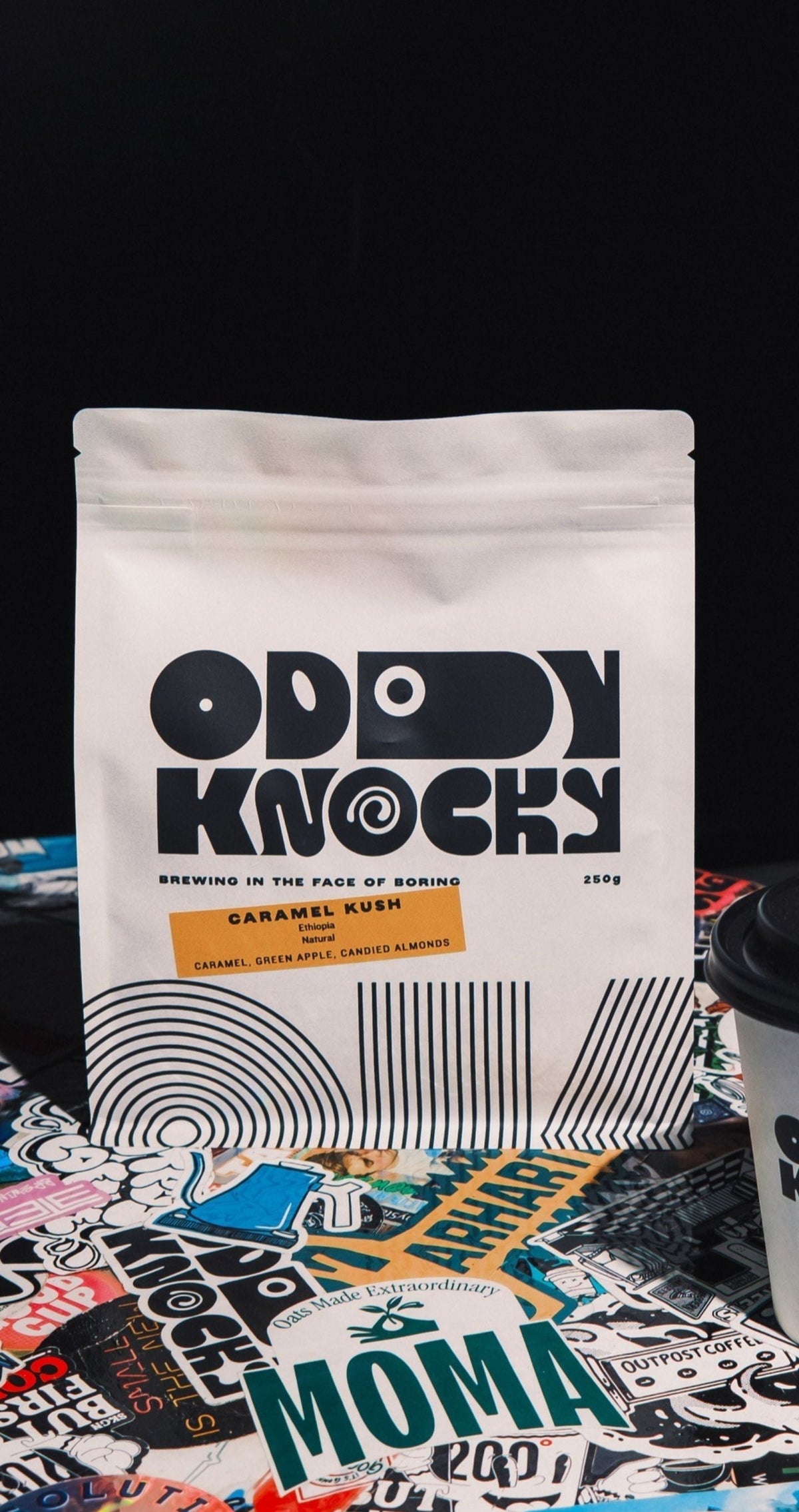 A white stand-up pouch of Oddy Knocky Caramel Coffee next to a white paper coffee cup with a black lid, both printed with the Oddly Knocky logo, set on a round table covered in colorful stickers against a black background.